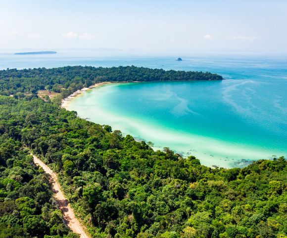 Aerial view of a lush green tropical forest bordering a turquoise bay with a sandy beach. The sky is clear, conveying a serene, idyllic atmosphere.