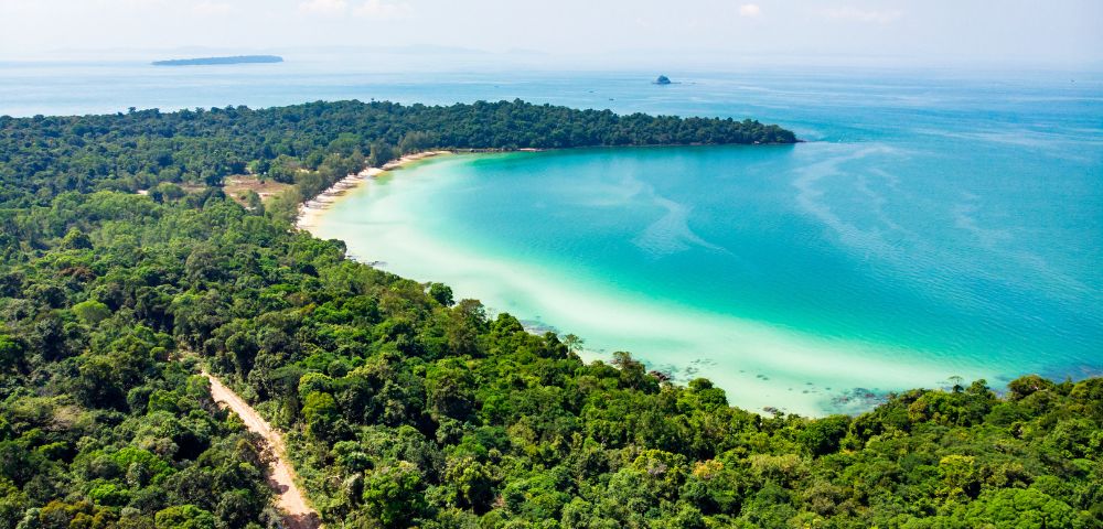Aerial view of a lush green tropical forest bordering a turquoise bay with a sandy beach. The sky is clear, conveying a serene, idyllic atmosphere.