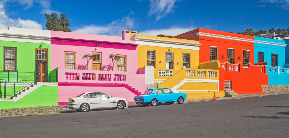 A row of vibrant houses in bright green, pink, yellow, orange, and blue with ornate windows. Two cars parked in front enhance the lively street scene.