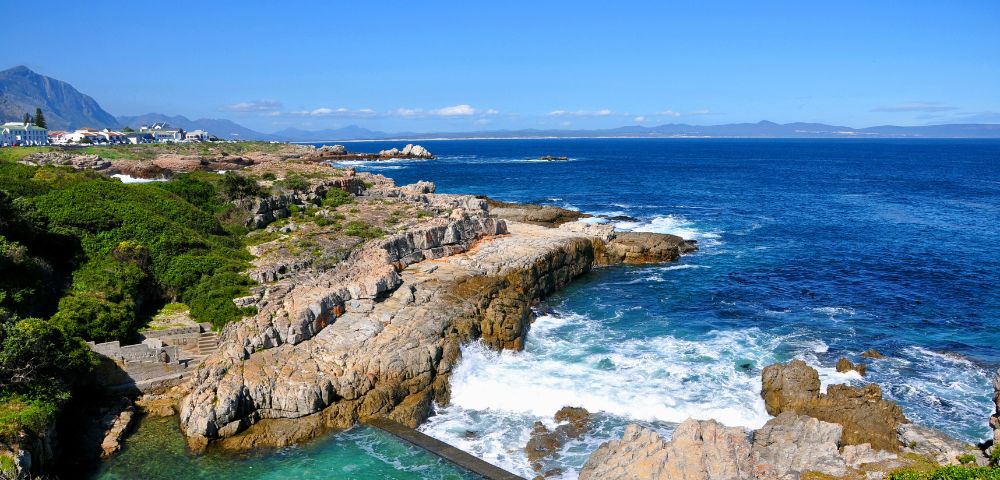Rocky coastline with green foliage, waves crashing against the shore under a clear blue sky. Distant mountains and a small town are visible, evoking tranquility.