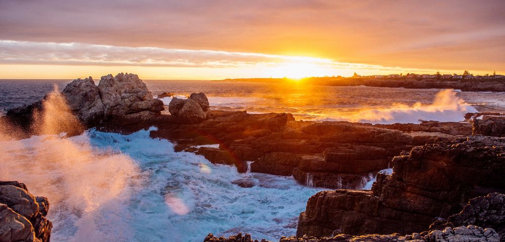 Sunset over rocky coastline with waves crashing against cliffs, creating a dynamic and vibrant scene. The sky is painted with warm orange and gold hues.