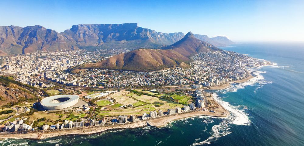 Aerial view of Cape Town with green hills, the distinctive circular stadium, urban buildings, and the coastline stretching alongside the bright blue ocean.