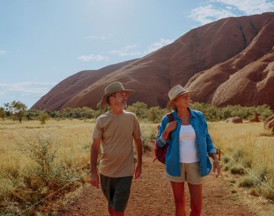 A man and woman in hats walk on a dirt path in a desert landscape, with a large rocky hill in the background under a clear, sunny sky.