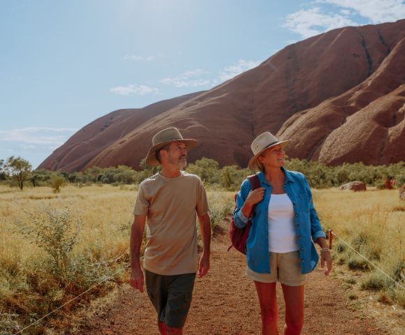 A man and woman in hats walk on a dirt path in a desert landscape, with a large rocky hill in the background under a clear, sunny sky.