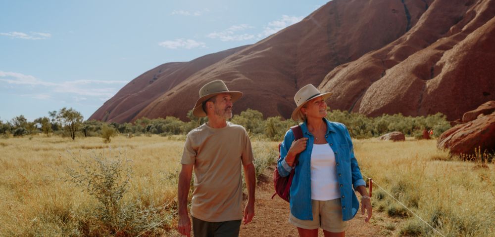 A man and woman in hats walk on a dirt path in a desert landscape, with a large rocky hill in the background under a clear, sunny sky.