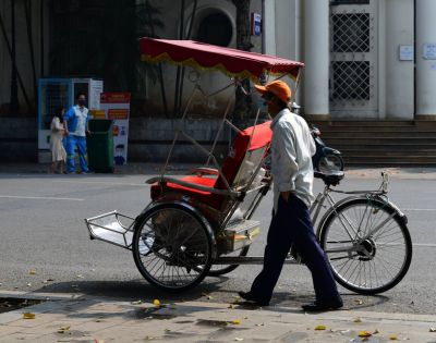 A rickshaw driver in a white shirt and orange cap walks beside his red and white rickshaw on a quiet urban street, conveying a calm, sunny day.
