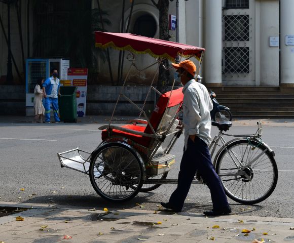 A rickshaw driver in a white shirt and orange cap walks beside his red and white rickshaw on a quiet urban street, conveying a calm, sunny day.