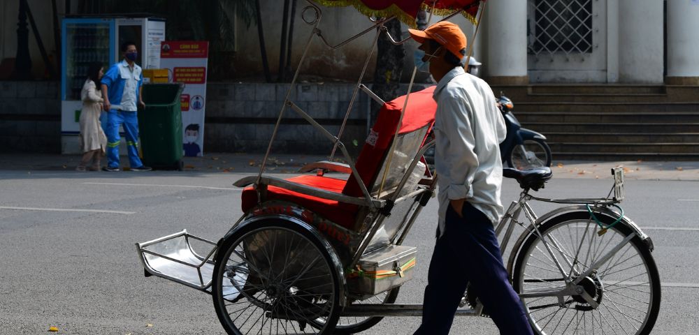 A rickshaw driver in a white shirt and orange cap walks beside his red and white rickshaw on a quiet urban street, conveying a calm, sunny day.