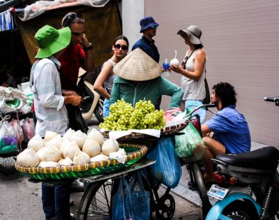 A street vendor in a conical hat sells coconuts and grapes from a bicycle cart. A group of people, casually dressed in hats and sunglasses, engage nearby.