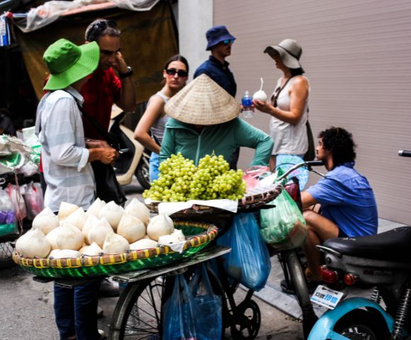A street vendor in a conical hat sells coconuts and grapes from a bicycle cart. A group of people, casually dressed in hats and sunglasses, engage nearby.