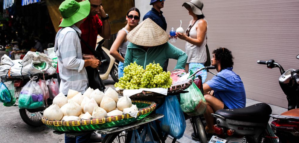 A street vendor in a conical hat sells coconuts and grapes from a bicycle cart. A group of people, casually dressed in hats and sunglasses, engage nearby.