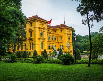 Bright yellow colonial-style building with a red roof, surrounded by lush green gardens. A red flag is flying on the roof, conveying a sense of grandeur.