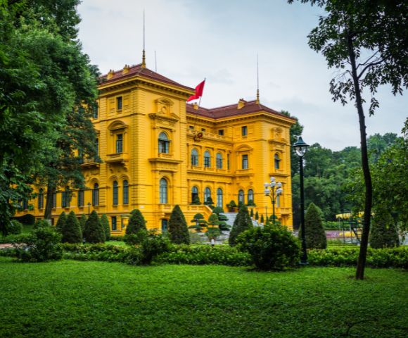 Bright yellow colonial-style building with a red roof, surrounded by lush green gardens. A red flag is flying on the roof, conveying a sense of grandeur.