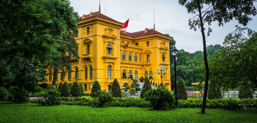 Bright yellow colonial-style building with a red roof, surrounded by lush green gardens. A red flag is flying on the roof, conveying a sense of grandeur.
