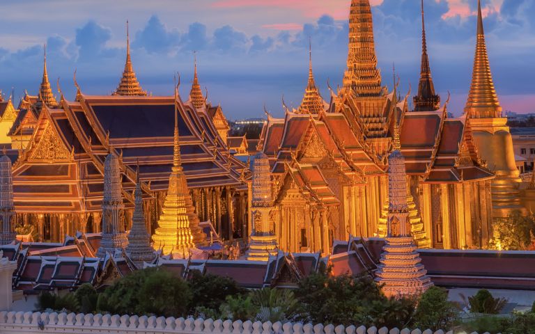 Intricate golden temple spires of the Grand Palace in Bangkok at dusk, glowing warmly against a vibrant sky with scattered clouds. Serene ambiance.