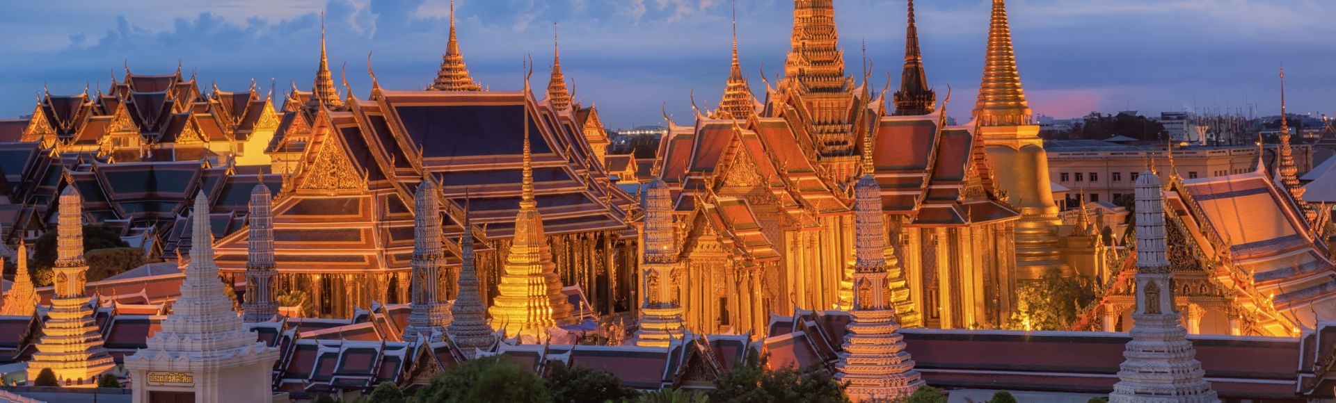 Intricate golden temple spires of the Grand Palace in Bangkok at dusk, glowing warmly against a vibrant sky with scattered clouds. Serene ambiance.