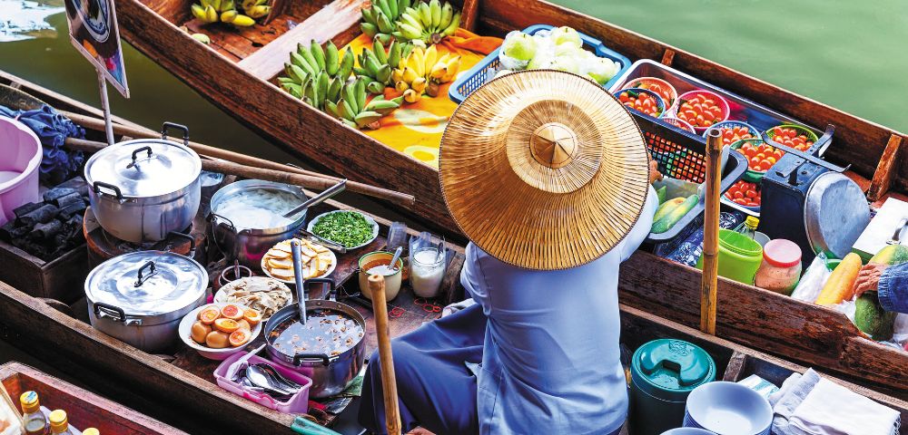 A vendor in a straw hat sits on a colorful boat at a floating market, surrounded by fruit, cooked food, and cooking utensils, creating a vibrant, lively scene.