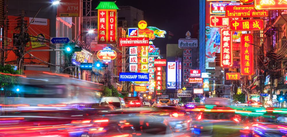 Bustling nighttime street in Chinatown, Bangkok filled with vibrant neon signs in various languages, and blurred car lights conveying energy and movement.