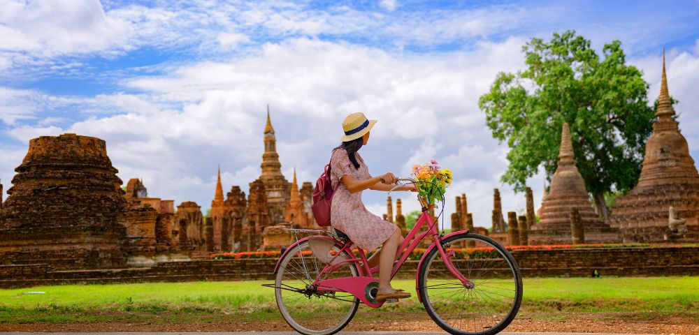 A woman on a pink bicycle, wearing a hat and dress, rides past ancient temples under a bright blue sky, conveying a sense of adventure and tranquility.