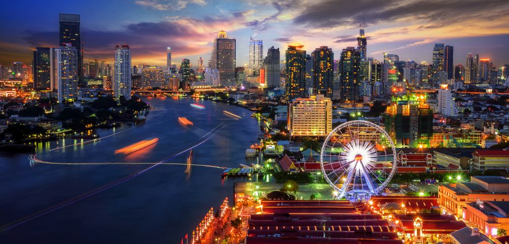 A vibrant cityscape at twilight with a glowing Ferris wheel by a river, skyscrapers illuminated against a colorful sky, creating a lively, energetic atmosphere.