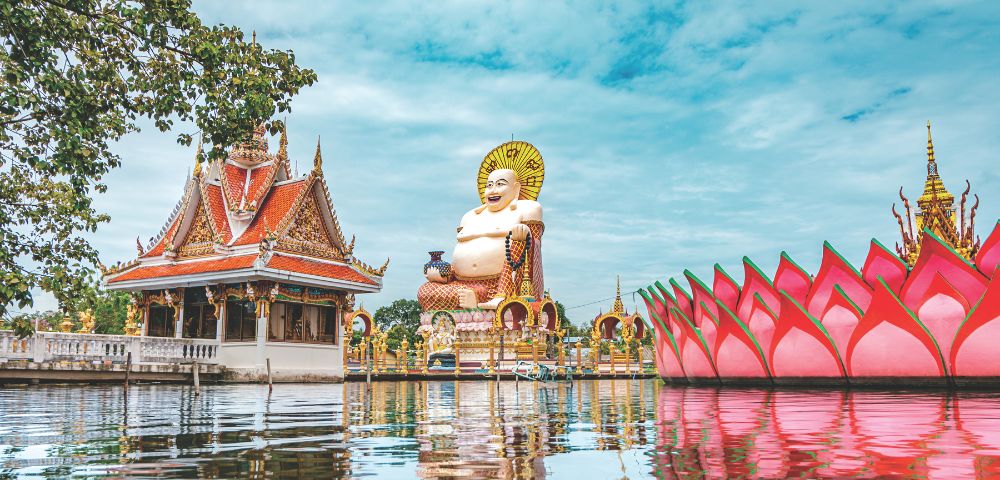 The image shows a serene temple scene with a large, smiling Buddha statue in the center. A pink lotus structure and ornate temple sit on calm water under a clear sky.
