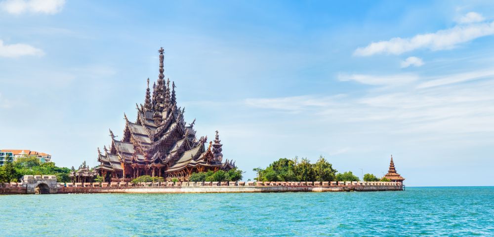 Ornate wooden temple with intricate spires sits on a waterfront, surrounded by greenery. Clear sky and turquoise water create a serene, majestic scene.