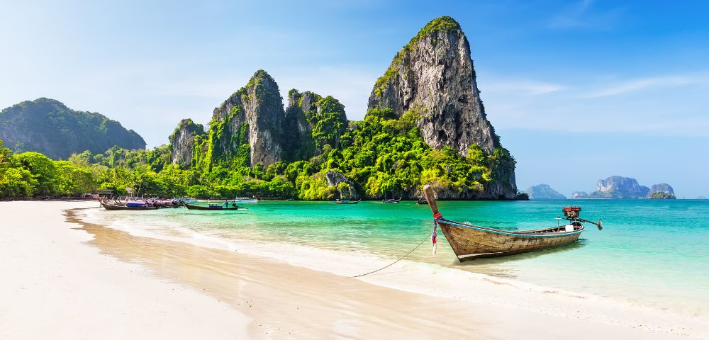 Serene tropical beach with white sand, turquoise water, and traditional long-tail boats. Tall limestone cliffs covered in greenery under a clear blue sky.