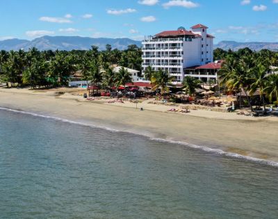 Aerial view of a serene tropical beach with a tall white hotel surrounded by palm trees. The calm ocean meets sandy shores under a clear blue sky.