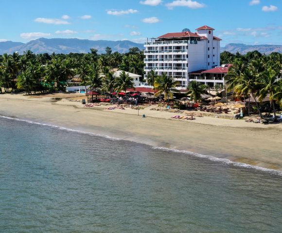 Aerial view of a serene tropical beach with a tall white hotel surrounded by palm trees. The calm ocean meets sandy shores under a clear blue sky.