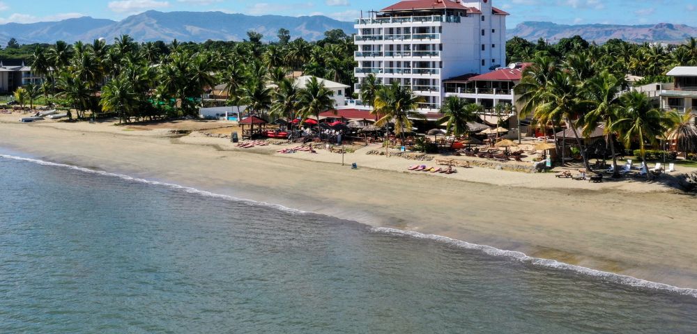 Aerial view of a serene tropical beach with a tall white hotel surrounded by palm trees. The calm ocean meets sandy shores under a clear blue sky.