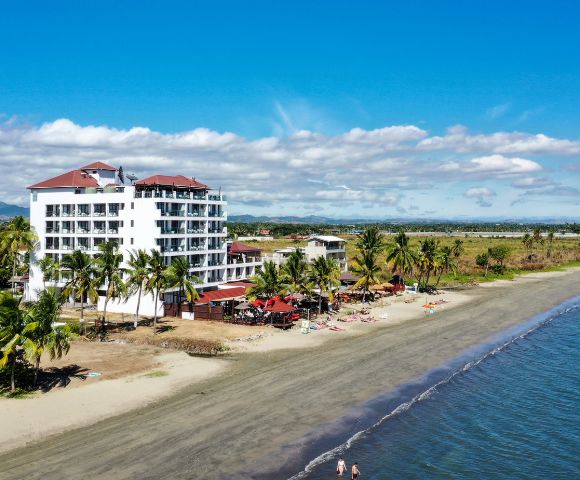 Aerial view of a beachfront hotel with red roofs, surrounded by palm trees. Relaxed beachgoers and vibrant umbrellas line the sandy shore under a clear blue sky.