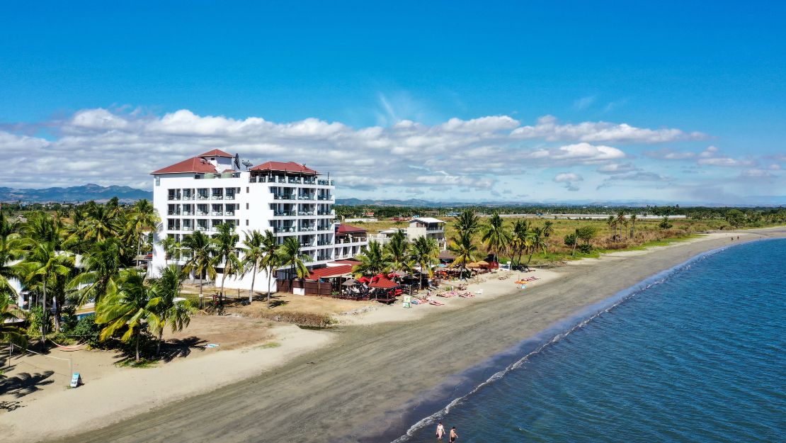 Aerial view of a beachfront hotel with red roofs, surrounded by palm trees. Relaxed beachgoers and vibrant umbrellas line the sandy shore under a clear blue sky.