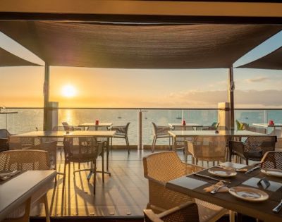 Beachfront restaurant with tables and chairs on a wooden deck, overlooking a serene ocean at sunset. Warm light creates a cozy and inviting atmosphere.
