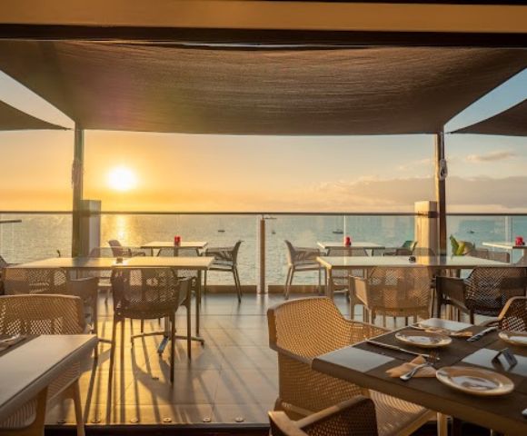 Beachfront restaurant with tables and chairs on a wooden deck, overlooking a serene ocean at sunset. Warm light creates a cozy and inviting atmosphere.