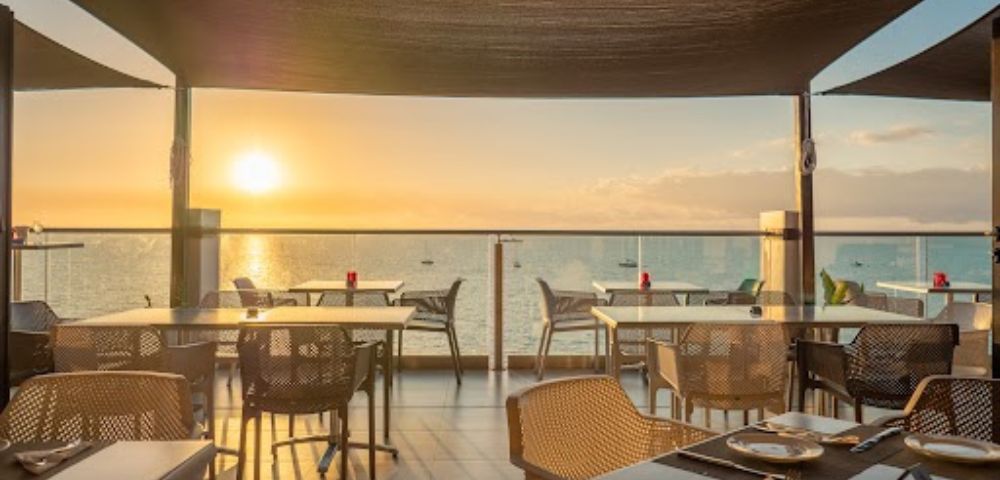 Beachfront restaurant with tables and chairs on a wooden deck, overlooking a serene ocean at sunset. Warm light creates a cozy and inviting atmosphere.
