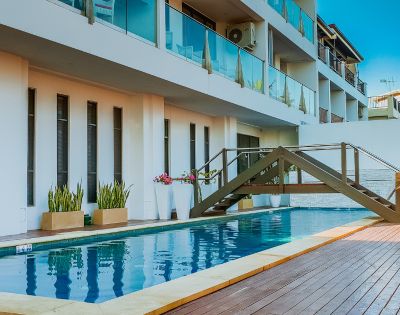 Modern hotel building with glass balconies overlooks a serene pool. A small wooden bridge crosses over, accentuating the relaxing atmosphere.