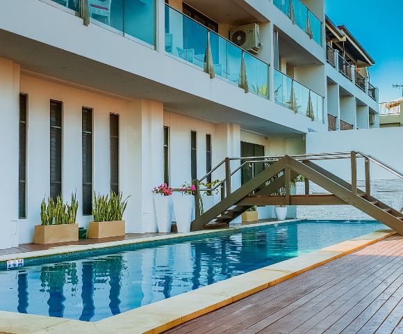 Modern hotel building with glass balconies overlooks a serene pool. A small wooden bridge crosses over, accentuating the relaxing atmosphere.