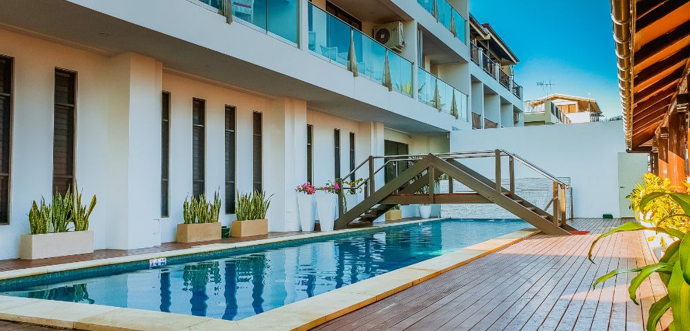 Modern hotel building with glass balconies overlooks a serene pool. A small wooden bridge crosses over, accentuating the relaxing atmosphere.