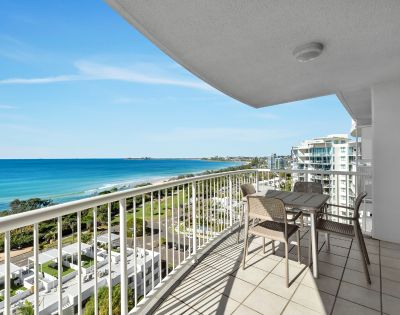 Spacious balcony overlooking a serene ocean view with blue skies, modern buildings, and green landscape. Set with a table and chairs, evoking relaxation.
