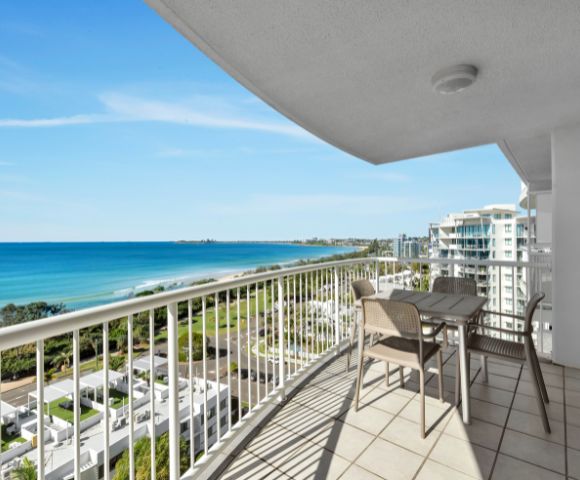 Spacious balcony overlooking a serene ocean view with blue skies, modern buildings, and green landscape. Set with a table and chairs, evoking relaxation.