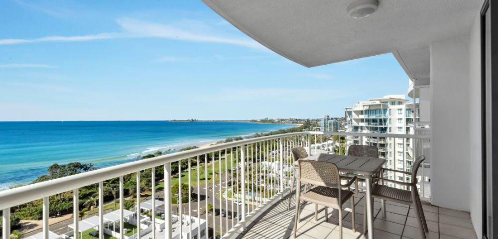 Spacious balcony overlooking a serene ocean view with blue skies, modern buildings, and green landscape. Set with a table and chairs, evoking relaxation.