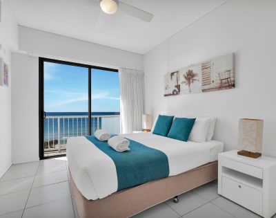 Bright, modern bedroom with a large bed, teal accents, and coastal artwork. Glass doors offer a stunning ocean view, creating a serene, airy atmosphere.