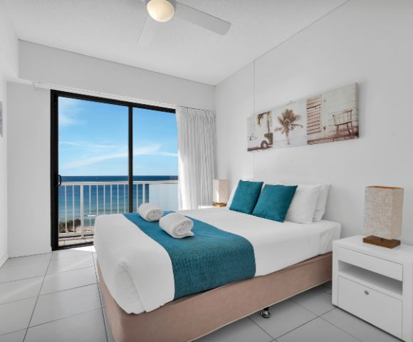 Bright, modern bedroom with a large bed, teal accents, and coastal artwork. Glass doors offer a stunning ocean view, creating a serene, airy atmosphere.