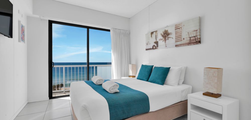 Bright, modern bedroom with a large bed, teal accents, and coastal artwork. Glass doors offer a stunning ocean view, creating a serene, airy atmosphere.