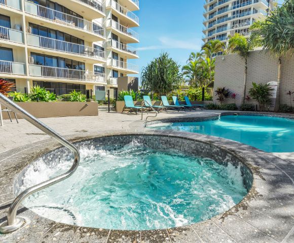 Hotel courtyard with a bubbling hot tub in the foreground and a turquoise swimming pool. Surrounded by lounge chairs and tropical plants under a clear sky.