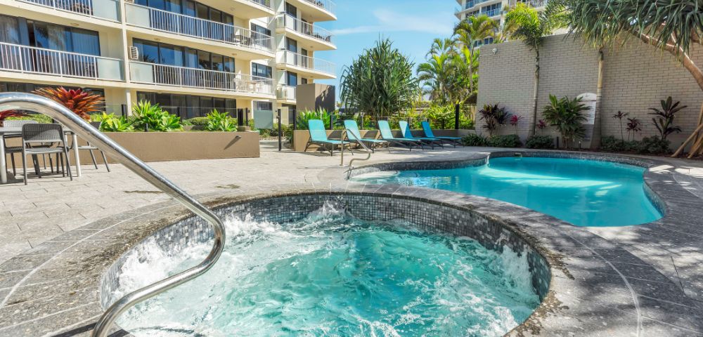 Hotel courtyard with a bubbling hot tub in the foreground and a turquoise swimming pool. Surrounded by lounge chairs and tropical plants under a clear sky.