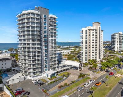 Aerial view of a modern beachfront urban area with two tall, sleek apartment buildings near the ocean under a clear blue sky. Cars are parked below.