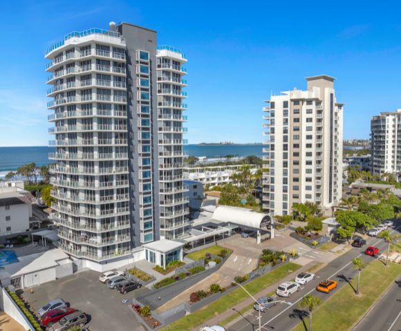 Aerial view of a modern beachfront urban area with two tall, sleek apartment buildings near the ocean under a clear blue sky. Cars are parked below.