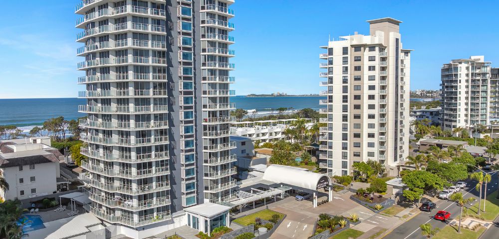 Aerial view of a modern beachfront urban area with two tall, sleek apartment buildings near the ocean under a clear blue sky. Cars are parked below.
