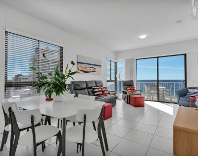 Bright, modern living room with ocean view. Features white walls, black sofas, red and blue accents, greenery, and glass doors leading to a sunny balcony.
